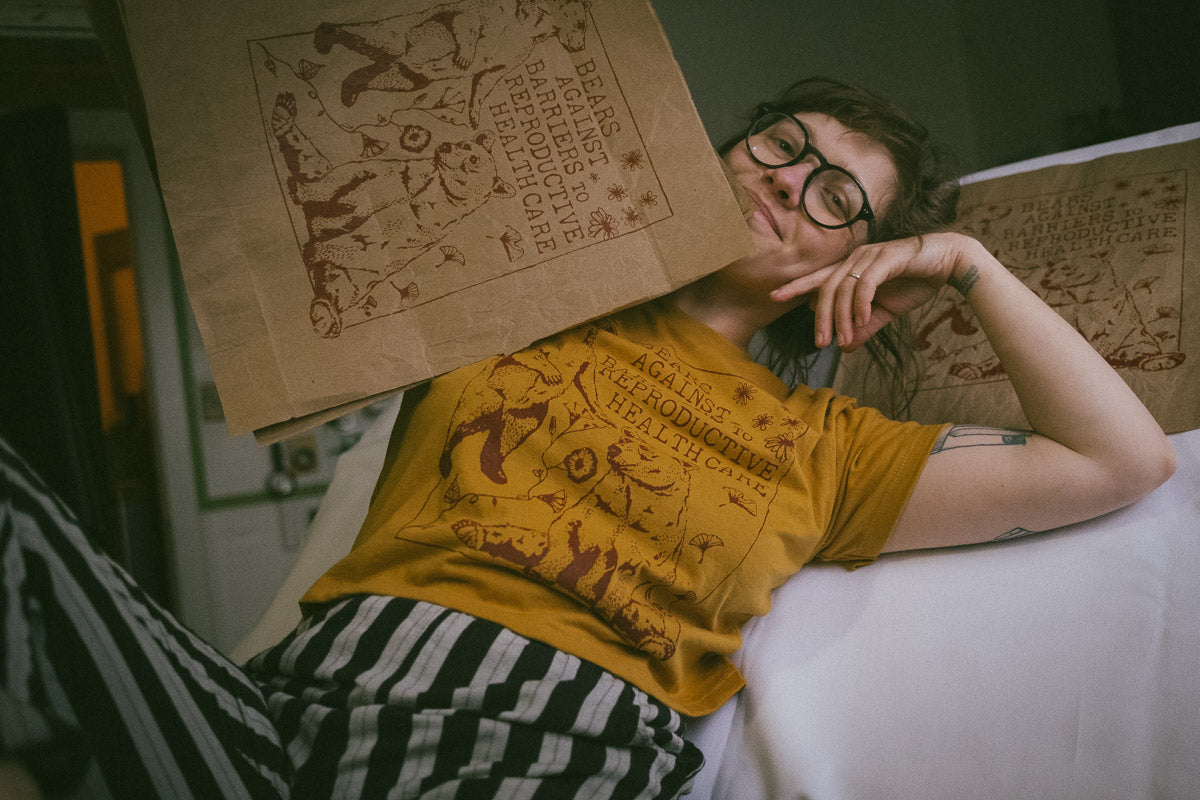 a woman with round glasses wearing the mustard yellow cropped feminist tee shirt holding the print on reused recycled kraft paper. she is wearing grey and black striped cotton pants from ace and jig