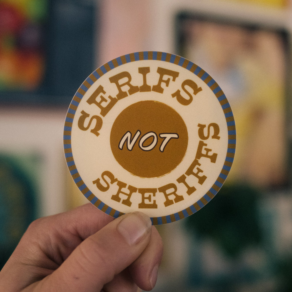 a hand holding a 3 inch circular sticker with a serif western typeface that reads "serifs not sheriffs" it has a bullseye design with a southwestern colorway