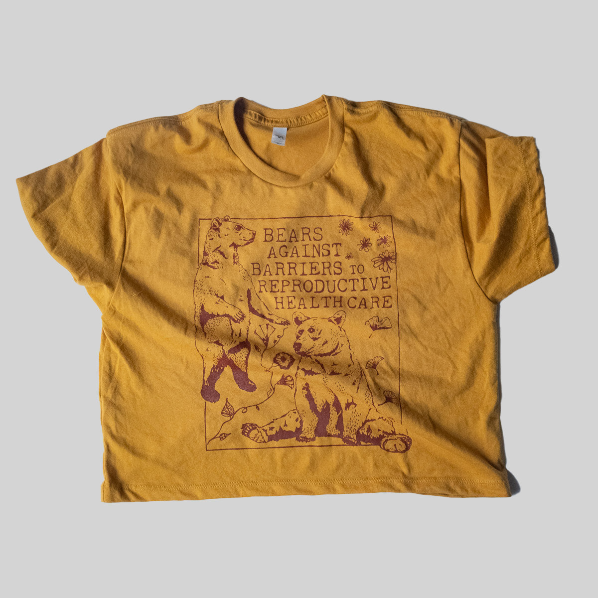 mustard yellow cropped tee with bears against barriers to reproductive health care illustrated screen print with red ink. drawing of flowers and gingko leaves. on a plain light grey background